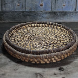 May include: A round, woven basket with a spiral design in the center. The basket is made of natural fibers in shades of brown and tan. The rim is a darker brown. The basket is approximately 10 inches in diameter.