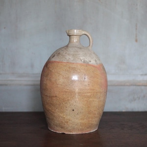 Op de afbeelding: Een antieke steengoed kruik met een handvat. De kruik heeft een afgeronde body en een smalle hals. De kleur is een mix van bruin, beige en een oranje band. Het wordt waarschijnlijk gebruikt voor het bewaren van vloeistoffen.