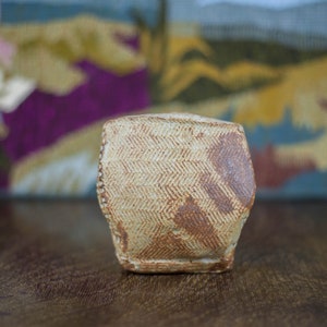 May include: A handmade ceramic tumbler with a textured, herringbone pattern in tan and brown. The small, hexagonal cup is displayed on a wooden surface, with a colorful, abstract tapestry in the background.