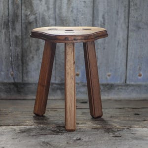 May include: A handcrafted wooden stool with three legs. The stool has a light-coloured, curved seat with three small holes. The legs and frame are made of a darker wood. The stool is approximately 46 cm tall.