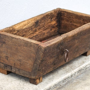 Peut inclure: Une jardinière en bois rustique de forme rectangulaire et de couleur brun foncé. La jardinière a un design simple avec quatre petites pattes et un anneau métallique sur le côté.