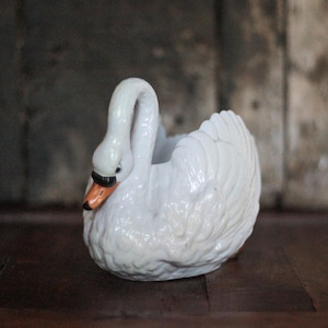 Peut inclure: Un pot de fleurs en forme de cygne en céramique blanche, avec un bec noir et des accents orange. Le cygne présente des textures de plumes détaillées et un cou incurvé, créant une pièce décorative et élégante.