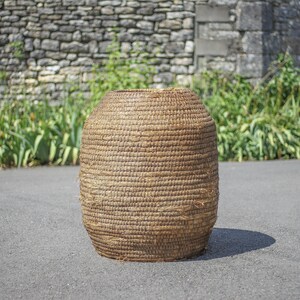 Peut inclure: Un grand panier rond en osier, fait de matériaux naturels. Le panier est brun et a une texture rugueuse. Il est posé sur une surface d'asphalte grise.