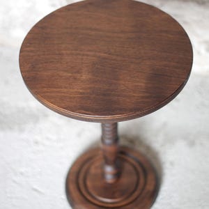 Pode incluir: Uma mesa de pedestal de madeira castanho-escura com uma superf&iacute;cie superior circular lisa. A mesa &eacute; suportada por uma coluna central &uacute;nica com um design torneado decorativo, assente numa base circular robusta com cor e acabamento a condizer.