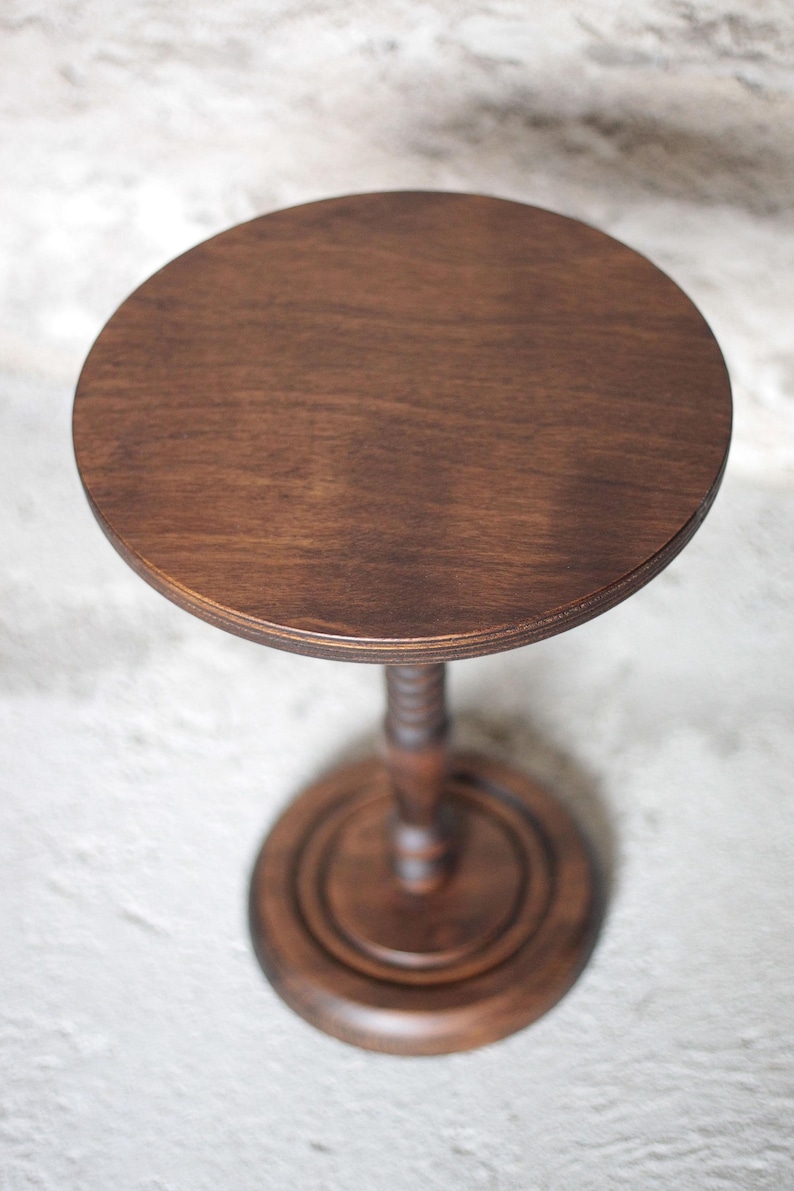 Pode incluir: Uma mesa de pedestal de madeira castanho-escura com uma superf&iacute;cie superior circular lisa. A mesa &eacute; suportada por uma coluna central &uacute;nica com um design torneado decorativo, assente numa base circular robusta com cor e acabamento a condizer.