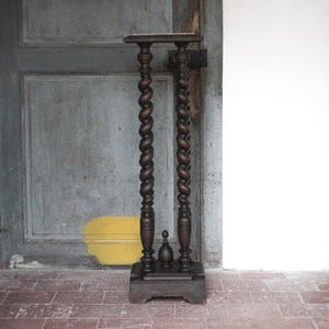 Peut inclure: Un présentoir à plantes antique, brun foncé, avec deux colonnes torsadées. Il possède une petite base carrée et une petite étagère. Le présentoir en bois est placé devant une porte grise. Hauteur d'environ 100 cm.