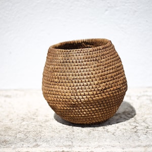 May include: A round, brown woven basket made of natural materials. The basket is empty and has a smooth, slightly textured surface.