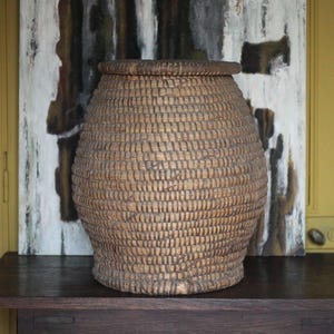 May include: A woven basket with a lid, made of natural-colored fibers. The basket has a rounded shape with a slightly wider base and a textured surface. It sits on a dark wooden table against a backdrop of abstract art and a yellow door.