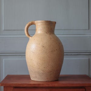 May include: A beige ceramic pitcher with a rounded body, a narrow neck, and a single curved handle on the side. The surface of the pitcher has a textured, speckled appearance with visible variations in the light tan colour throughout the exterior.