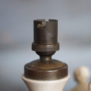 May include: Close-up of a vintage lamp socket. The socket is made of brass and is mounted on a white ceramic base. The brass socket has a cylindrical shape with a slot for the light bulb. The ceramic base has a textured surface.