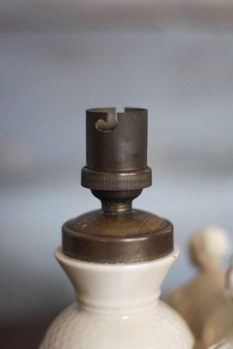 May include: Close-up of a vintage lamp socket. The socket is made of brass and is mounted on a white ceramic base. The brass socket has a cylindrical shape with a slot for the light bulb. The ceramic base has a textured surface.