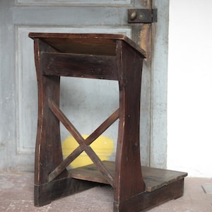 Peut inclure: Un prie-dieu en bois brun foncé avec un dessus incliné, une petite étagère et un repose-pieds. Le prie-dieu a un support en forme de X sur les côtés et est en bois vieilli. Fond avec une porte grise et un sol carrelé rouge.