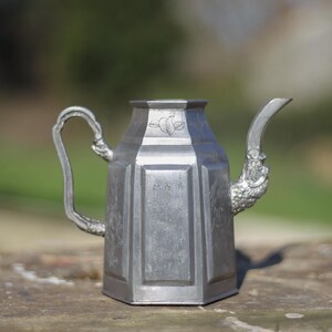 Peut inclure: Une théière en argent avec une poignée décorative et un bec verseur. La théière a un corps carré avec un sommet arrondi et une surface texturée.