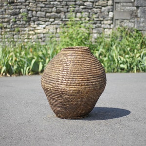 May include: A large, round, woven basket made of natural materials. The basket is brown and has a slightly textured surface. It is sitting on a gray asphalt surface.