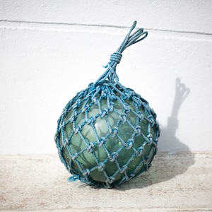 Può includere: Una boa di vetro blu con una rete di corda blu. La boa è rotonda e ha uno sfondo bianco.