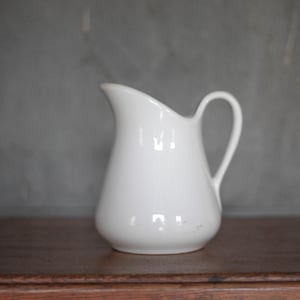 Op de afbeelding: Een witte keramische kan met een rond lichaam en een handvat. De kan staat op een houten oppervlak.