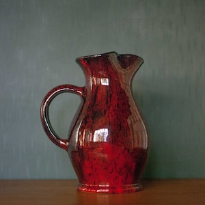 Könnte beinhalten: Eine rote Keramikkanne mit einem gesprenkelten Überzug. Die Kanne hat einen breiten Boden und einen schmalen Hals mit einem Ausguss. Der Griff ist an der Seite der Kanne befestigt.