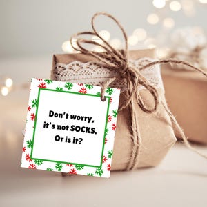 May include: A white gift tag with a green and red snowflake pattern. The tag has a green border and the text "Don't worry, it's not SOCKS. Or is it?" in black.