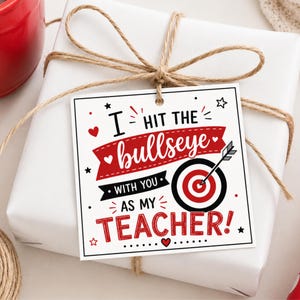 May include: A white gift box tied with twine, featuring a gift tag with the text "I hit the bullseye with you as my teacher!" The tag has a target design with a red bullseye and an arrow.