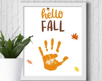 Puede incluir: Una impresión enmarcada en blanco con el texto "hello fall" en letras marrones y naranjas. Una huella de mano naranja está en el centro de la impresión con pequeñas ilustraciones de hojas naranjas y amarillas alrededor.