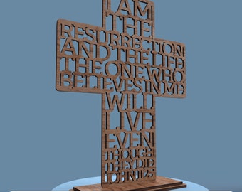 May include: A wooden cross with the words "I AM THE RESURRECTION AND THE LIFE THE ONE WHO BELIEVES IN ME WILL LIVE EVEN THOUGH THEY DIE JOHN 11:25" cut out. The cross is on a wooden base. The bottom of the image reads "DIGITAL FILE FOR LASER CUTTING".