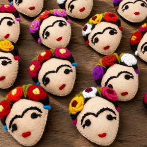 May include: A collection of twelve felt appliques, each shaped like a face with embroidered features including thick eyebrows, dark eyes, and red lips. Each face is adorned with a crown of small, colorful felt flowers in red, yellow, and purple.