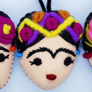 May include: Three felt ornaments shaped like faces with black hair, embroidered floral headpieces in bright colors including pink, yellow, purple, and orange, thick black eyebrows, closed eyes with eyelashes, and small red stitched mouths.