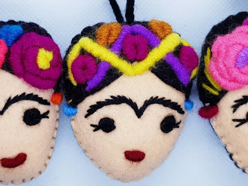 May include: Three felt ornaments shaped like faces with black hair, embroidered floral headpieces in bright colors including pink, yellow, purple, and orange, thick black eyebrows, closed eyes with eyelashes, and small red stitched mouths.