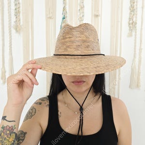 May include: A wide-brimmed, natural-toned straw hat with a black band and chin strap. The hat has a classic fedora shape and is suitable for sun protection. The hat is a neutral color and can be worn by anyone.