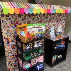 May include: A display booth with a colorful striped awning and floral patterned backdrop featuring a black metal shelving unit filled with various toys, including an archery set, a wooden chest, a blue football, and a second black shelf unit.