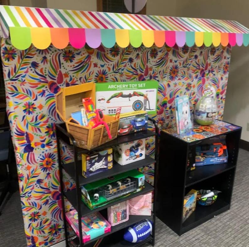 May include: A display booth with a colorful striped awning and floral patterned backdrop featuring a black metal shelving unit filled with various toys, including an archery set, a wooden chest, a blue football, and a second black shelf unit.