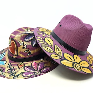 Può includere: Due cappelli fedora viola con fasce nere. I cappelli presentano disegni dipinti a mano che includono fiori gialli, foglie verdi, un cuore rosso e un uccellino blu. La trama intrecciata dei cappelli è visibile e la vernice è applicata in modo grafico.