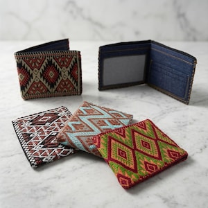 May include: Four bifold wallets featuring woven fabric exteriors with geometric patterns in red, black, white, blue, and green. One wallet is open, revealing a dark blue denim interior with a clear ID window and card slots, while three are folded.