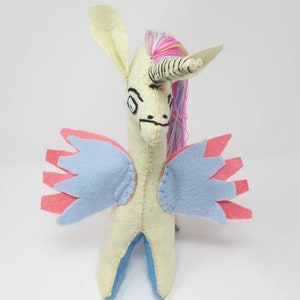 Puede incluir: Un peluche de unicornio de fieltro hecho a mano con cuerpo color crema, cuerno a rayas y crin de hilo multicolor. La criatura tiene alas superpuestas de color azul claro y rosa, rasgos faciales cosidos y patas interiores azules, de pie.