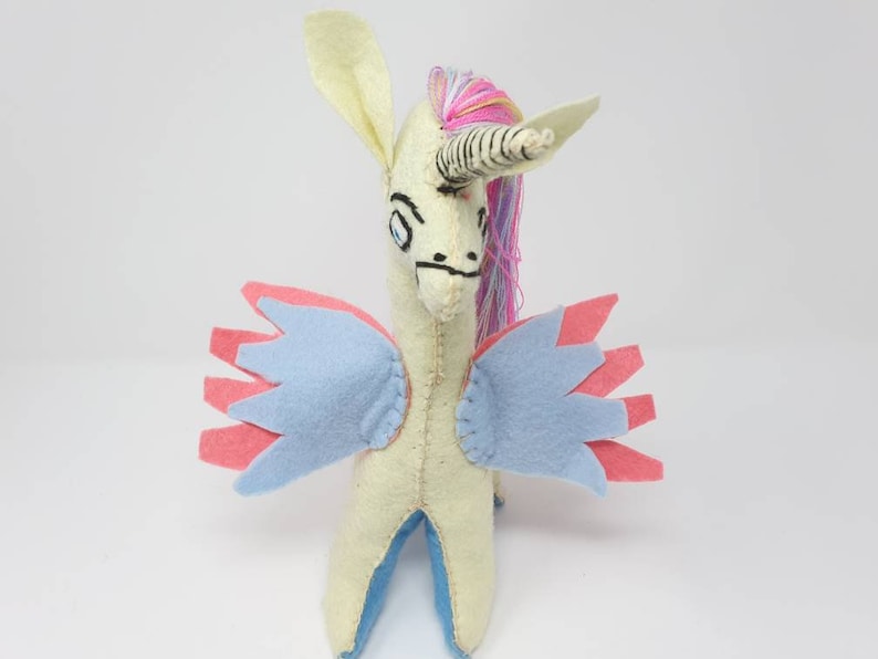 Puede incluir: Un peluche de unicornio de fieltro hecho a mano con cuerpo color crema, cuerno a rayas y crin de hilo multicolor. La criatura tiene alas superpuestas de color azul claro y rosa, rasgos faciales cosidos y patas interiores azules, de pie.