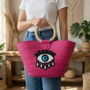 May include: A vibrant fuchsia woven straw tote bag featuring a circular light-coloured braided handle and a prominent embroidered graphic of a stylised eye with black lashes and a blue iris centred on the front of the bag.