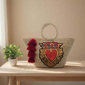 May include: A woven straw tote bag featuring a circular handle and a large, central embroidered patch of a red heart with a gold sequined border and a blue and red crown. A tiered red tassel hangs from the left side of the bag's front panel.