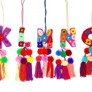 May include: Five colourful felt letter ornaments with pom-pom tassels. The letters are K, L, M, N, and O. Each letter is embroidered with colourful flowers and has a pink, purple, red, or green tassel.
