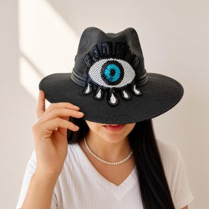 May include: A black fedora hat featuring a prominent embroidered eye design on the front. The eye motif is crafted with white, black, and bright blue sequins, including teardrop-shaped accents below the eye. The hat has a structured brim.