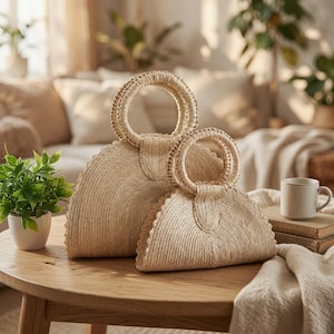 May include: Two semi-circular handbags crafted from woven natural straw fibers. Each bag features a rigid, circular handle attached to the top center. The bags are light beige in color and display a consistent, textured, braided straw pattern throughout.