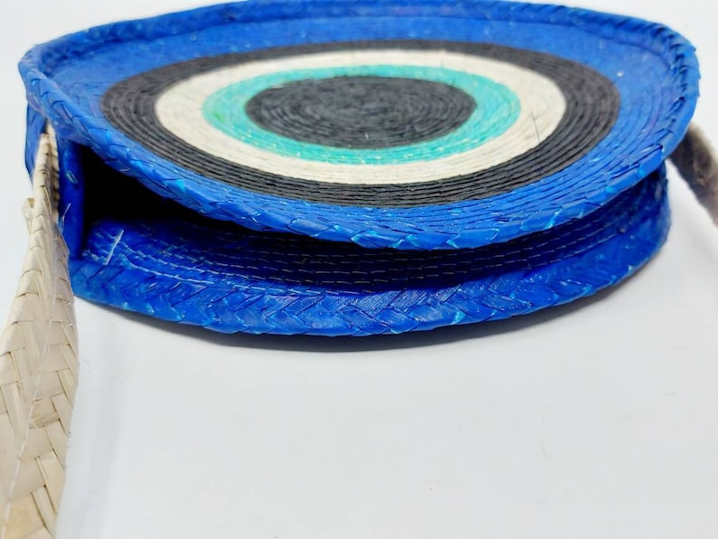 May include: A round, blue and white woven bag with a white woven strap. The bag has a circular pattern of blue, white, and black.