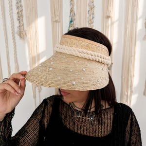 May include: A straw visor featuring a wide, natural-coloured woven brim adorned with scattered white pearl-like beads. The crown of the visor is wrapped with a thick, cream-coloured twisted rope trim that encircles the base of the headpiece.