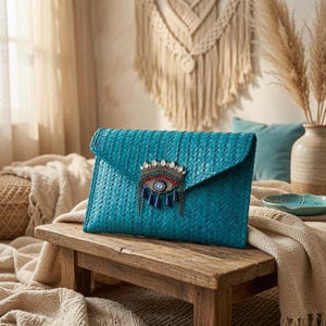 Puede incluir: Un bolso de mano tipo sobre tejido en color verde azulado con un adorno decorativo de ojo turco en la solapa frontal. El adorno está hecho de metal plateado, cristales transparentes, cuentas rojas y azules, y colgantes en forma de gota.