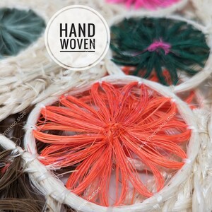 Pu&ograve; includere: Un tessuto intrecciato a mano con motivi circolari a fibre radianti nei toni dell'arancione, verde scuro e marrone, su uno sfondo intrecciato beige naturale. Una sovrapposizione grafica circolare bianca contiene il testo nero HAND WOVEN.