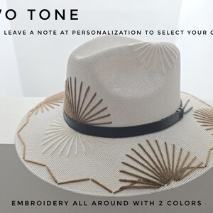 May include: A cream-colored fedora hat featuring a black leather band and sunburst-style embroidery in two shades of brown. The text on the image reads: TWO TONE, PLEASE LEAVE A NOTE AT PERSONALIZATION TO SELECT YOUR COLORS, EMBROIDERY ALL AROUND WITH 2 COLORS.