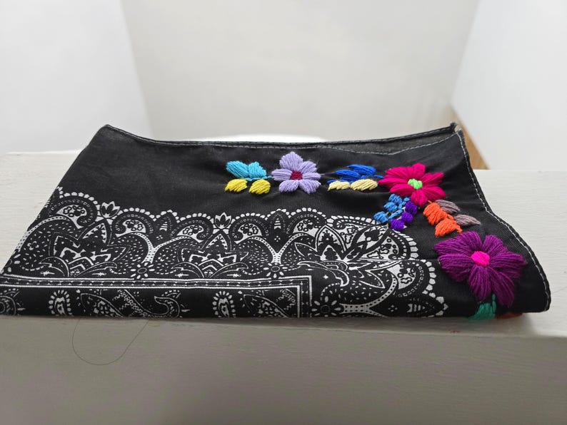 May include: A black bandana featuring a white paisley print along the bottom edge and colorful floral embroidery in shades of pink, purple, blue, yellow, and orange stitched onto the upper right corner of the fabric.