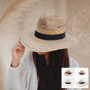 May include: A light brown straw fedora hat featuring a wide brim, a pinched crown, and a solid black grosgrain ribbon band. The hat includes three small metal eyelets on each side of the crown for ventilation, showcasing a natural woven texture.