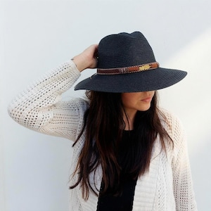Peut inclure: Un chapeau fedora gris anthracite foncé avec un large bord et un tissage texturé semblable à de la paille. Le chapeau présente une bande en cuir marron ornée de petites perles métalliques et d'une breloque plume dorée sur le devant.
