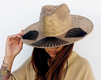 Puede incluir: Un sombrero fedora de paja color canela con una corona pellizcada y un ala ancha. El sombrero está decorado con tres patrones de bordado negros en forma de abanico cosidos en el ala, que irradian desde la base de la corona en un diseño.