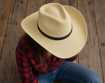 Puede incluir: Un sombrero de vaquero de color beige claro con una copa estructurada y un ala ancha y curvada. El sombrero presenta una banda negra sencilla y lisa que rodea la base de la copa, creando un contraste limpio con el material texturizado.